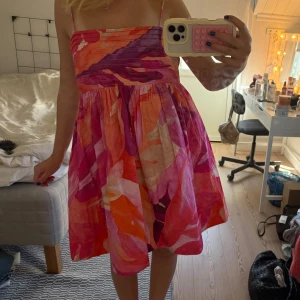 Färgglad kort sommarklänning - Superfin kort klänning med tunna axelband och färgstarkt abstrakt mönster i rosa, orange, rött och lila toner. Klänningen har en rynkad överdel och utställd kjol, perfekt för varma dagar. Materialet känns lätt och luftigt.