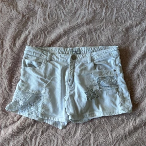 Vita spetsdetaljerade shorts Mayoral - Snygga vita shorts från Mayoral med broderade spetsdetaljer och blommiga inslag på fickor och ben. Går att dra åt med resår i midjan. Knappen har rostat lite men inget som syns.