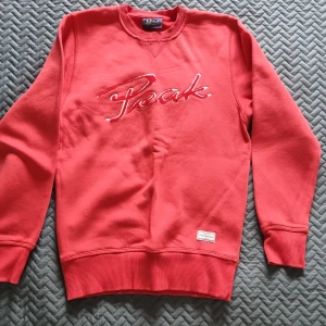 Röd Peak Performance sweatshirt - Säljer en röd sweatshirt från Peak Performance med broderad logga framtill. Tröjan har rund halsringning, ribbade muddar och är tillverkad i mjuk bomull. Perfekt för dig som gillar en sportig och avslappnad stil.