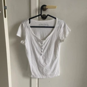 Vit kortärmad topp med knappar - En basic vit topp från Kappahl med korta ärmar och v-ringning. Framsidan har en rad små knappar som ger en snygg detalj. Toppen är i mjuk bomull och har en enkel, clean look som passar till allt.