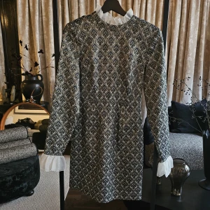 Custommade klänning - Rosetta - Unik kortklänning med svart, silver och guldigt mönster, långärmad och med vit volangkrage samt transparenta volangdetaljer vid ärmslut. Baksidan har en stor rosett i vitt tyg och en öppen detalj. Perfekt för dig som vill sticka ut med en elegant och cool look.