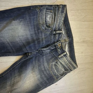 Blå jeans med broderade detaljer - Snygga blå jeans från Kaporal med slitna partier och vita broderade detaljer på fickorna. Jeansen har raka ben, klassisk femficksmodell och coola nitar vid fickorna. Perfekt för dig som gillar en unik och trendig look.