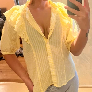 Söt blus med fina detaljer - Ljus gul blus med vita ränder och volanger på axlarna samt fina detaljer vid kragen. Perfekt till sommaren. Som man ser på bilderna kan man ha den på sig precis som man vill, vilket kan förändra looken helt. Vill man ha den lite öppnare kan man knäppa upp 2-3 knappar, eller så kan man bära den helt stängd som jag har på en av bilderna. Köpt på Lindex för 299 kr 2022, använt kanske 2 ggr, den har endast tagit plats i garderoben pga blusar inte riktigt är min stil. Men helt underbar blus alltså!