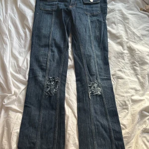 Blå bootcut jeans med fickor och slitningar - Snygga blå jeans med bootcut passform och coola slitningar lite nedanför knäna. Byxorna har dubbla knappar i midjan och två framfickor med lock och silverfärgade knappar. Klassisk jeanslook med markerade sömmar och bälteshällor.