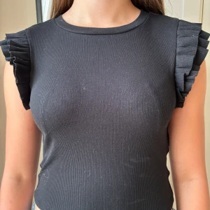 Svart topp med volangärm  - Supersnygg svart ribbad topp från Zara med volangdetaljer på axlarna. Säljer på grund av att den tyvärr inte passar mig. Pris kan diskuteras!!❣️
