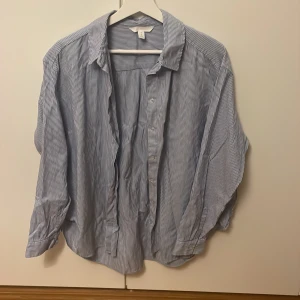 Blåvit randig skjorta från H&M, M - Klassisk blåvit randig skjorta från H&M i storlek M. Skjortan har lång ärm, knappar framtill och en avslappnad passform. Tillverkad i mjuk bomull och passar perfekt till jeans eller chinos. Snygg krage och diskreta vita knappar ger en clean look.