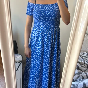 Blå blommig offshoulder maxiklänning  - Supersöt blå långklänning från new york med vita småblommor, offshoulder och smockad överdel. Klänningen har korta ärmar med knyt och ett flowy fall. Perfekt för sommarens alla tillfällen, riktigt skön och luftig känsla.