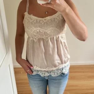 Beige spetslinne i satin - Supersött beige linne med tunna axelband och vacker spets upptill och nertill. Tillverkat i glansig satin med romantiska spetsdetaljer som ger en feminin vibe. Perfekt att styla med jeans eller kjol för en chic look.