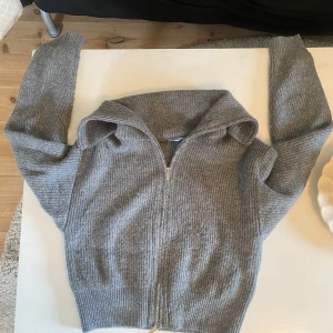 Grå ribbad kofta från NA-KD - Mysig grå ribbstickad kofta från NA-KD i storlek XS. Koftan har lång ärm, dragkedja framtill och en bred krage. Perfekt för lager-på-lager och chill dagar. Snygg och enkel att matcha med jeans eller kjol.