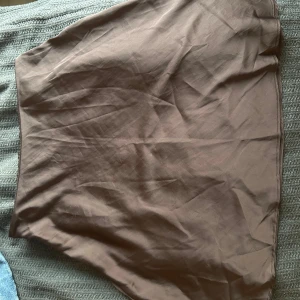 Brun kortkjol i satin - Enkel och stilren brun kortkjol med lätt glansig yta i satin material . Kjolen har en rak passform. Dragkedja i sidan för smidig påklädning.