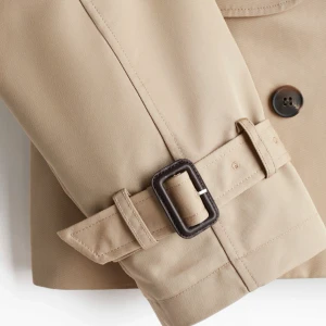 Kort beige trenchcoat  - Snygg croppad trenchcoat i beige med dubbelknäppning och stora slag. Jackan har bältdetaljer vid ärmsluten och klassisk krage.                  Lägger ut för att se intresse då den inte kommit till användning, slutsåld på hemsidan