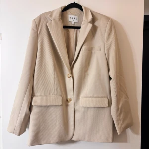 Beige kavaj från NA-KD - Stilren beige kavaj från NA-KD med klassisk krage, två knappar framtill och fickor med lock. Kavajen har en rak passform och är tillverkad i ett strukturerat tyg som ger en snygg look. 