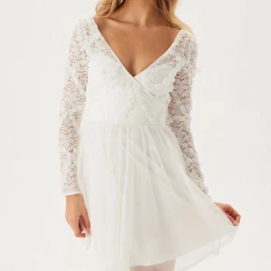 Vit spetsklänning med v-ringning - Superfin vit kortklänning med v-ringad omlottfront och långa spetsärmar. Perfekt som studentklänning! Helt oanvänd, är fortfarande kvar i paketet från när jag köpte den