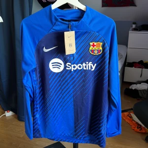 Blå långärmad FC Barcelona Nike-tröja - Säljer en blå långärmad FC Barcelona träningströja från Nike med Dri-FIT-material. Tröjan har klubbmärke, Nike-logga och Spotify som sponsor på bröstet. Snyggt diagonalt mönster framtill och dragkedja vid halsen. Perfekt för dig som älskar Barca och vill ha en stilren look. Helt ny, men var tyvärr för liten för mig. Storlek S