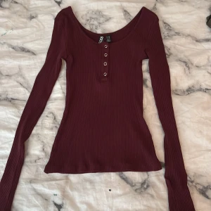 Vinröd ribbad långärmad topp från Pieces - Vinröd långärmad topp från Pieces i ribbad bomullsmix. Snygg v-ringning och fyra silvriga knappar framtill ger en clean look. Tight passform. Tröjan är i storlek xxs men materialet är väldigt stretchigt så det passar faktiskt som vanlig xs eller s, helt oanvänd 