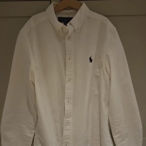 Vit skjorta från Ralph Lauren - Klassisk vit skjorta från Ralph Lauren med broderad logga på bröstet. Skjortan har lång ärm, knappar framtill och är tillverkad i mjuk bomull. Perfekt för en clean och stilren look.