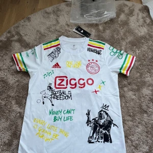 Ajax x Bob Marley Adidas fotbollströja - Ajax x Bob Marley limited edition fotbollströja från Adidas. Vit med färgglada ränder i grönt, gult och rött på axlar och ärmar. Tryck med Bob Marley, graffiti-inspirerade texter och Ajax-logga. Tillverkad i lätt och ventilerande Aeroready-material.