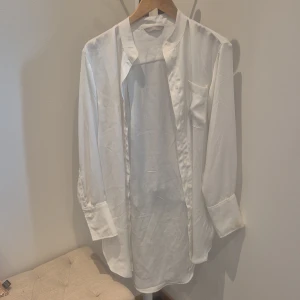 Vit långärmad skjorta från H&M - Vit långärmad skjorta från H&M med klassisk rak passform och ståkrage. Skjortan har bröstficka och knäppning framtill. Tillverkad i ett slätt och lätt material som ger en clean look. Perfekt för dig som gillar minimalistisk stil.