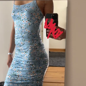 Blommig blå klänning från SHEIN - Ljusblå klänning från SHEIN med smala axelband och blommigt mönster i vitt, rött och grönt. Justerbar på sidan och stretchig.