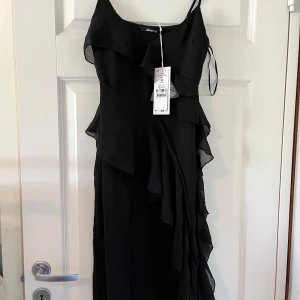 Svart långklänning med volanger från Gina Tricot - Supersnygg svart långklänning från Gina Tricot med tunna axelband och volangdetaljer längs sidorna. Klänningen har en elegant och flowy passform som ger en cool och trendig look. Perfekt för dig som vill sticka ut med stil.