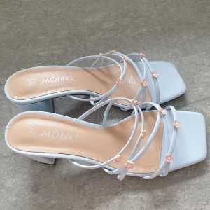 Blå sandaler med blommor från Monki - Snygga ljusblå sandaler från Monki med fyrkantig tå och blockklack. Remmarna är dekorerade med små rosa blommor för en söt och trendig look. Veganskt material och perfekt för dig som vill sticka ut med detaljer.