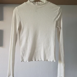Vit ribbad långärmad tröja  - Vit ribbad långärmad tröja från H&M Divided med hög hals och vågig kant vid halsringning och nederkant. Tight passform och stretchigt material som sitter snyggt. Perfekt att styla med jeans eller kjol för en clean look. Lite lätt genomskinlig (iaf om man har vit bh under). 