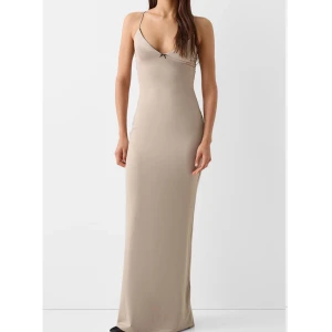 Beige långklänning från Zara - Supersnygg beige långklänning från Zara i figurnära modell med tunna axelband och v-ringning. Klänningen har en liten rosett framtill och är tillverkad i ett mjukt, stretchigt material som sitter perfekt på kroppen.