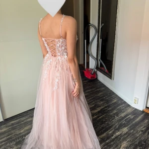 Ljusrosa långklänning med spetsdetaljer - Drömmig ljusrosa långklänning med tunna axelband, v-ringning och snörning i ryggen. Klänningen har vackra spets- och blomapplikationer upptill och ett svepande tyll-lager som ger en sagolik känsla. Perfekt för bal eller bröllopsgäster. Aldrig använd ute. Pris kan diskuteras. 