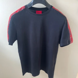 Svart t-shirt från Hugo med röda detaljer - Svart t-shirt från Hugo med röda band längs axlarna där det står HUGO. Klassisk passform, rund hals och korta ärmar. Tillverkad i mjuk bomull som känns skön mot huden. Perfekt för en clean och stilren look.