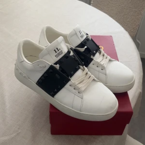 Valentino Rockstud vita sneakers - Snygga vita Valentino Rockstud sneakers i skinn med bred svart rem över vristen dekorerad med svarta nitar. Klassisk rund tå, vita snören och coola detaljer längs sulan. Perfekta för dig som vill sticka ut med en edgy men stilren look.