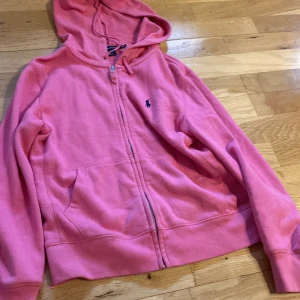 Rosa hoodie från Ralph Lauren - Rosa hoodie från Ralph Lauren med dragkedja framtill och klassisk logga broderad på bröstet. Tröjan har huva med snörning och två fickor framtill. Perfekt för en chill och avslappnad stil.