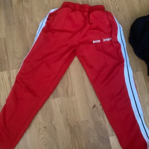 Palm Angels röda mjukisbyxor med ränder - Röda mjukisbyxor från Palm Angels med vita sidoränder och broderad logga på låret. Byxorna har elastisk midja och raka ben, tillverkade i mjukt syntetmaterial. Perfekta för en sportig och trendig streetwear-look.