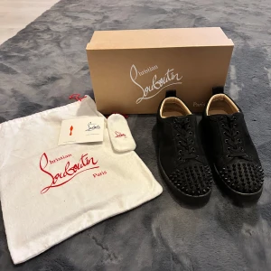 Christian Louboutin svarta sneakers med nitar - Svarta sneakers från Christian Louboutin i mocka med coola svarta nitar på tån och klassisk röd sula. Skorna har snörning och rund tå, och kommer med originalpåse och box. Perfekta för dig som vill sticka ut med en edgy vibe.