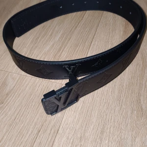 Svart skärp i skinn - Snyggt svart skärp i skinn med prägladeloggor längs hela bältet och en stor spänne i metall. Perfekt accessoar för att lyfta din outfit med en lyxig touch.