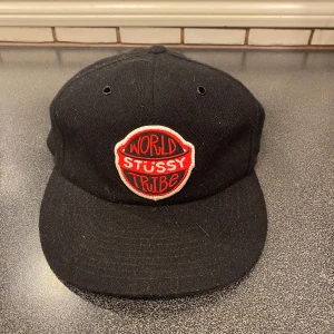 Svart Stussy keps - Riktigt fet svart keps från Stussy med broderad 'World Stussy Tribe'-patch i rött och vitt framtill. Inte mycket använd och säljes pga det. Sitter bra på huvudet och är justerbar baktill. Tveka inte på att höra av er om ni har frågor! Mvh Hampus 