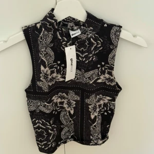 Svart mönstrad ärmlös topp Gina Tricot - Snygg svart ärmlös topp från Gina Tricot med hög hals och detaljerat paisleyinspirerat mönster i vitt. Croppad modell som sitter tajt och ger en cool vibe. Perfekt att styla med jeans eller kjol för en trendig look.