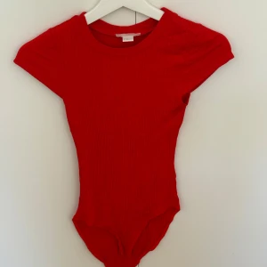 Röd ribbad bodysuit från H&M - Säljer en tight röd ribbad bodysuit från H&M i storlek XS. Bodyn har korta ärmar, rund halsringning och är tillverkad i stretchigt material som sitter snyggt på kroppen. Perfekt att styla med jeans eller kjol för en cool look. Testad nån gång