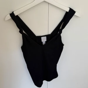 Svart ribbad linnetopp från H&M - Svart ribbad linnetopp från H&M i storlek XS. Toppen har breda axelband med snygga knutdetaljer och v-ringning framtill. Perfekt att styla med jeans eller kjol för en chill look. Materialet är mjukt och stretchigt för extra komfort.