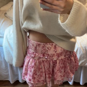 Rosa blommig kortkjol - Söt rosa kortkjol med blommigt mönster och volangdetaljer. Perfekt för en somrig look. Kjolen har en elastisk midja för bekväm passform.