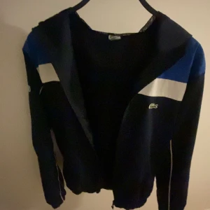 Mörkblå jacka från Lacoste - Säljer en riktigt snygg vindjacka från Lacoste. Jackan är i fint skick. Enda defekten är att en av de små dragkedjorna krånglar lite. Säljs då den är för liten för mig.