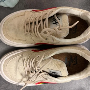 Beige sneakers med regnbågsdetalj från Eytys - Snygga beige sneakers från Eytys med en färgglad regnbågsdetalj på sidan. Skorna har snörning och en mörkblå rand på hälen. Perfekta för att ge en färgklick till din outfit. Skorna är i bra skick, lite smutsiga men utan andra defekter.