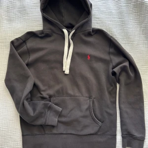 Svart hoodie från Ralph Lauren - Säljer en stilren svart hoodie från Ralph Lauren med det klassiska röda logotypen på bröstet. Hoodien har en justerbar dragsko i vitt och en praktisk magficka. Storlek M. om man vill kan man byta tillbaka till de vanliga svarta snören
