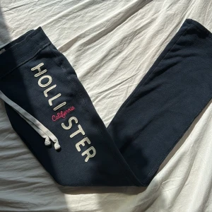 Mörkblåa  mjukisbyxor från Hollister - Så sjukt snygga lågmidjade mörkblåa mjukisbyxor från Hollister med vit snörning i midjan och texten 'Hollister California' längs benet. Byxorna har bootcut och sitter så snyggt på! Perfekta för en avslappnad stil. Jättegamla så går ej att få tag i längre. Midjemått tvärsöver: 33 cm. Innerbenslängd: 76 cm. Storlek XS.