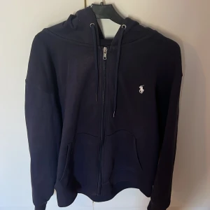 Svart hoodie från Ralph Lauren - Säljer en stilren svart hoodie från Ralph Lauren med dragkedja och det klassiska logotypbroderiet på bröstet. Tröjan har en justerbar huva med snören och praktiska fickor framtill. Perfekt för en avslappnad look.