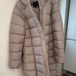 Beige dunjacka från 157 - Säljer en snygg beige dunjacka från 157. Jackan har en lång design med dragkedja och knappar framtill. Perfekt för kyliga dagar med sin vadderade stil och huva för extra värme. En stilren och praktisk jacka för vintern.