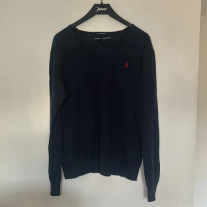 Mörkblå tröja från Ralph Lauren Sport - Säljer en snygg mörkblå tröja från Ralph Lauren Sport med en röd broderad logga på bröstet. Tröjan har en v-ringad design och långa ärmar, perfekt för en stilren look. Boyfriend fit för en avslappnad passform. Inga fläckar. Nästan som ny. 