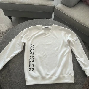 Vit sweatshirt från Moncler - Säljer en stilren vit sweatshirt från Moncler med svart logotyptryck längs sidan. Tröjan har långa ärmar och en rund halsringning. Perfekt för en avslappnad och trendig look.