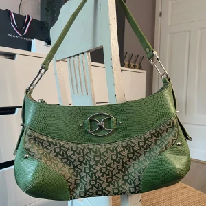 Grön axelväska från DKNY - Snygg grön axelväska från DKNY med ett unikt mönster och metallspänne framtill. Väskan har en dragkedja upptill och är perfekt för att bära dina dagliga nödvändigheter. Den har en  axelrem och är tillverkad i syntetmaterial.