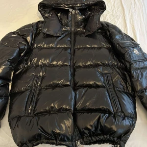 Svart pufferjacka från Moncler - Snygg svart pufferjacka från Moncler med glansig finish. Jackan har en dragkedja framtill och praktiska fickor med dragkedjor. Den har en justerbar huva och ett unikt serietidningsmönster på insidan. Perfekt för kyliga dagar!
