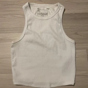 Vit ribbad linnetopp från FSBN Sister - Säljer en stilren vit ribbad linnetopp från FSBN Sister. Perfekt för varma dagar med sin ärmlösa design och bekväma passform. Toppen är enkel att matcha med olika outfits. 